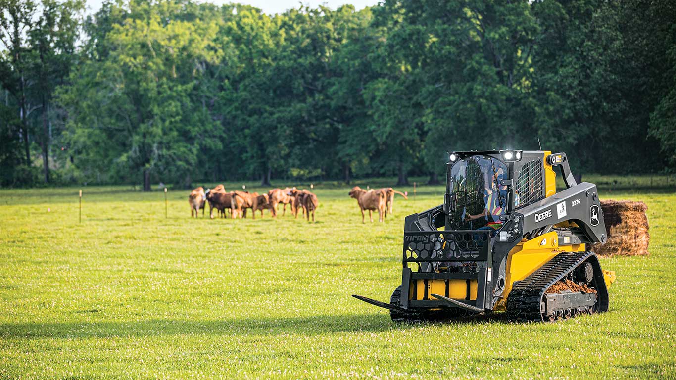 333G Skid Steer with bale spears in a field with cattle and a hay bale in the background