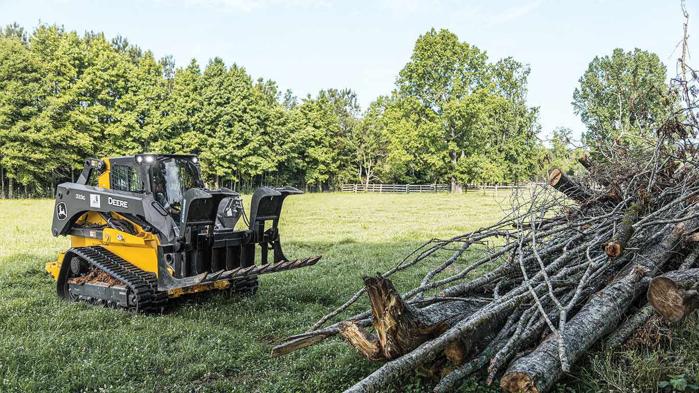 333G Skid Steer with a grapple attachment next to tree trunks in a grass field with trees in the distance