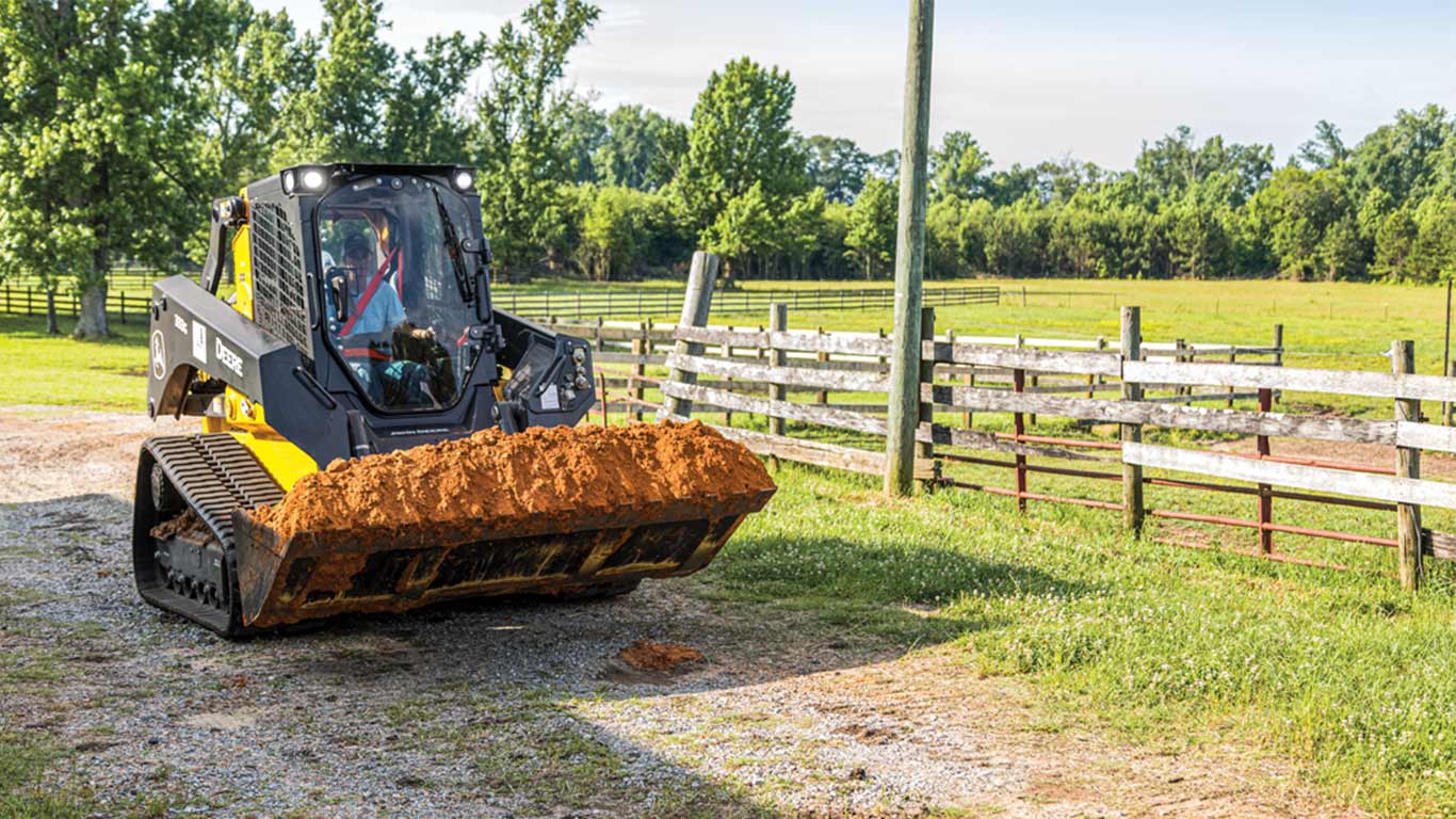 330 G-Tier Skid Steer with a loader attachment full of orange dirt alongside a wooden fence with a field and trees in the background