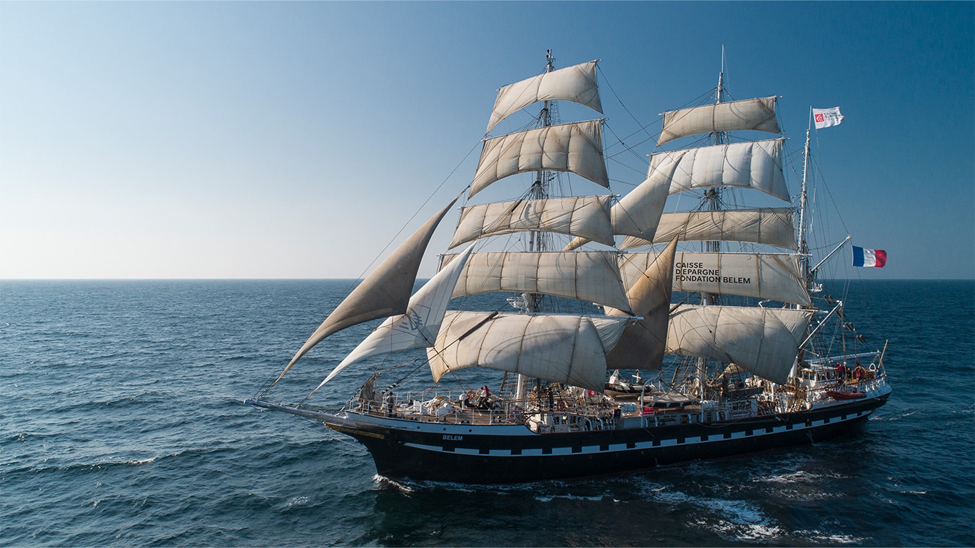 A large three-masted tall ship with white sails and a French flag flying from the mast sails on the ocean.