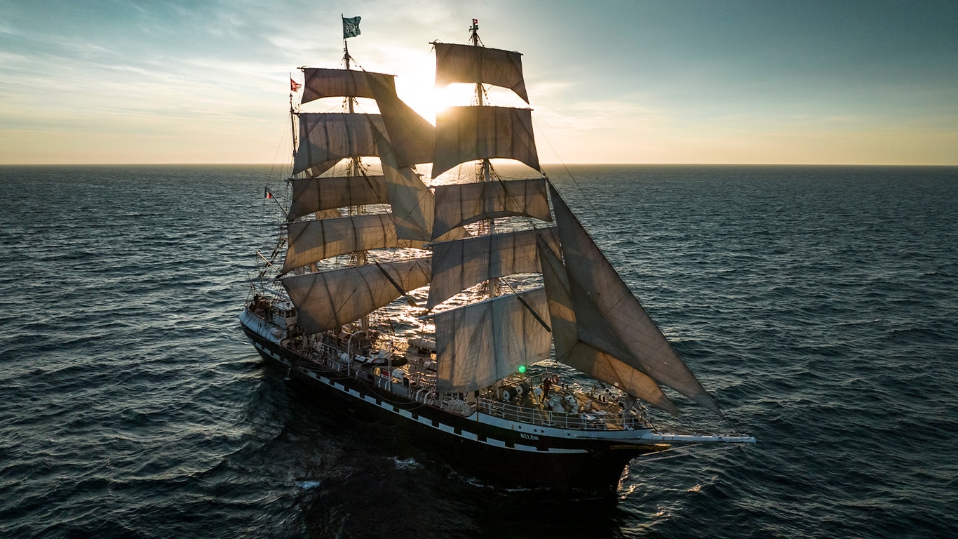 A large three-masted tall ship with white sails backlit by the setting sun.