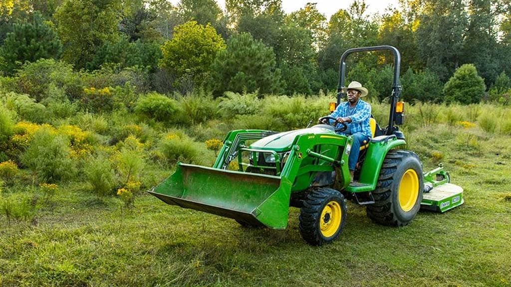 A person in a cowboy hat drives a green compact tractor with a bucket on the front and a mowing implement being pulled.
