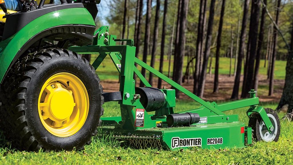 Close up of a green rotary cutter cutting grass in front of a tree line.