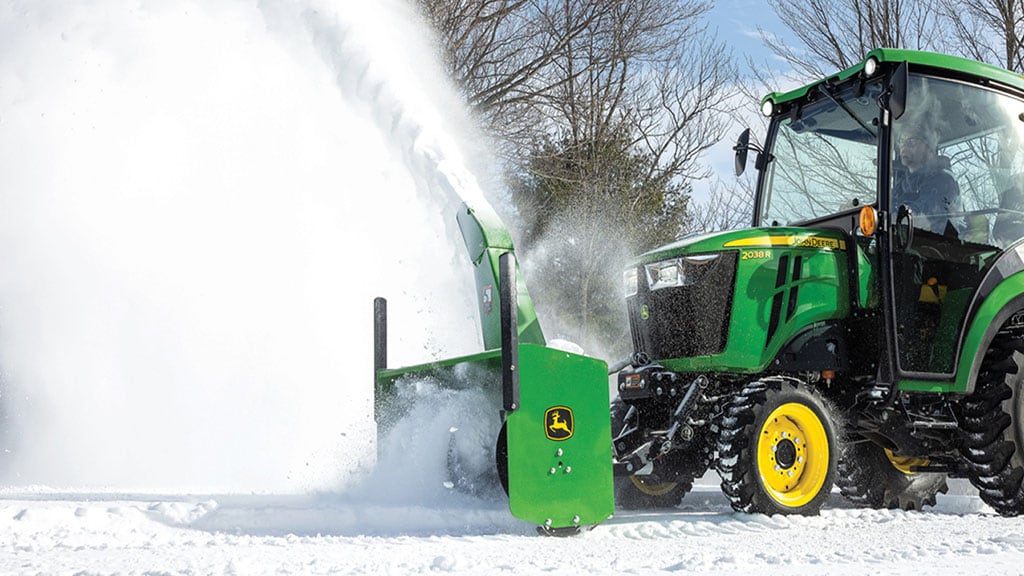 A green cabbed tractor blowing snow with a snow removal implement.