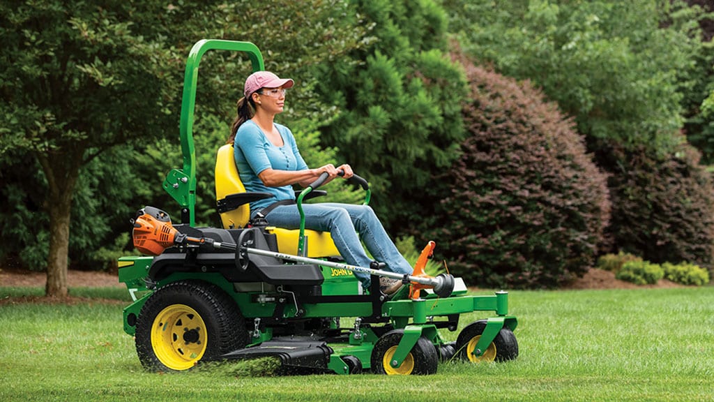 A person in safety glasses mowing a well-landscaped lawn in a green zero-turn mower.
