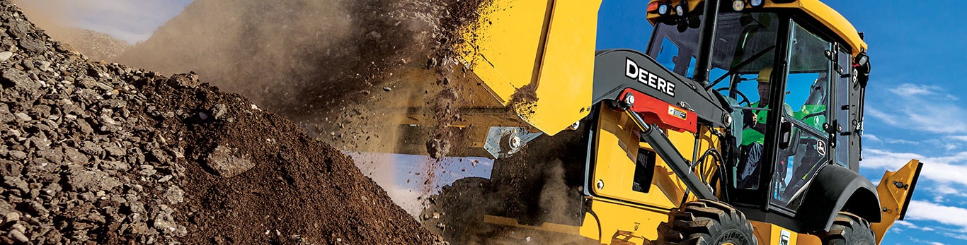 A John Deere backhoe unloads a scoop of dirt onto a pile