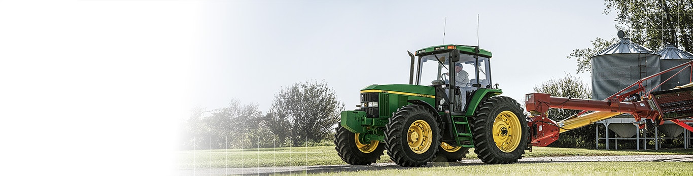 A John Deere tractor towing a red implement past grain bins on a farm.