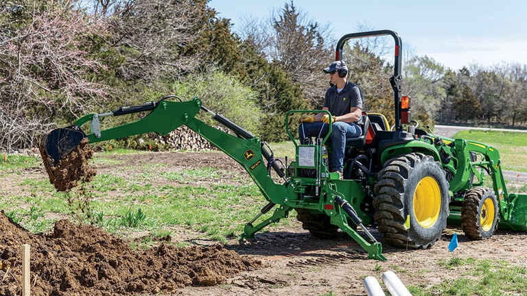 A person with ear and eye protection using a green backhoe to dump dirt in a pile (9th thumbnail)