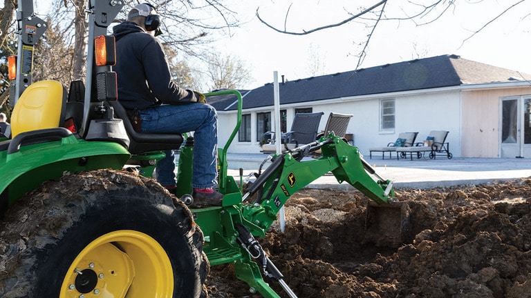 A person with ear and eye protection using a green backhoe to dump dirt in a pile (10th thumbnail)
