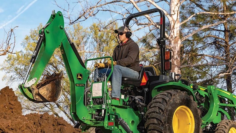 A person with ear and eye protection using a green backhoe to scoop soil. (8th thumbnail)