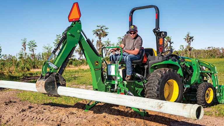 A green backhoe attached to a green compact tractor holds a large pipe above a trench. (12th thumbnail)