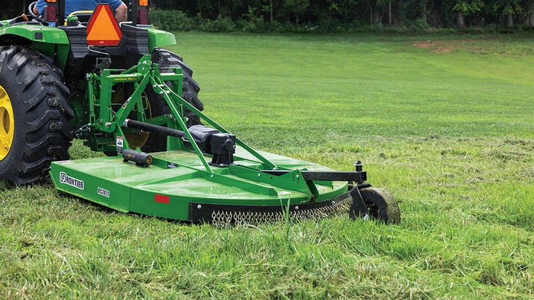A green rotary cutter attached to the end of a compact tractor evening up a large area of grass.