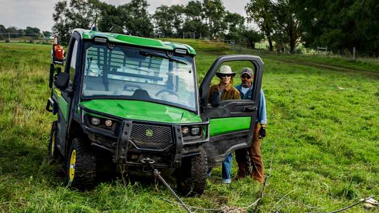 Two individuals stand behind an open driver-side door of a cabbed green Gator while a taut chain is attached to the front. (10th thumbnail)