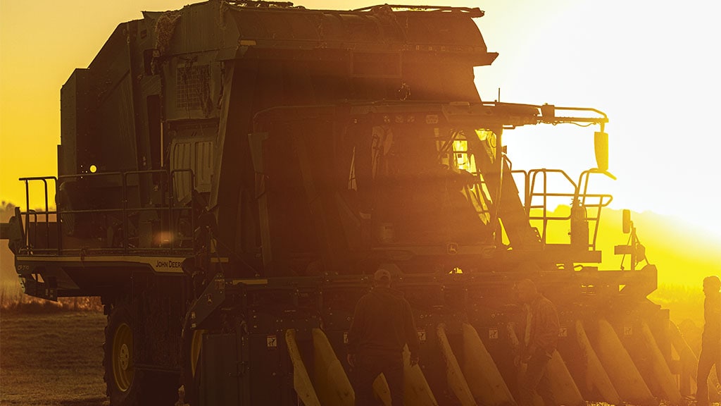 Front view of a CP770 Cotton Picker in a dusty field at dusk, with the setting sun behind the harvester