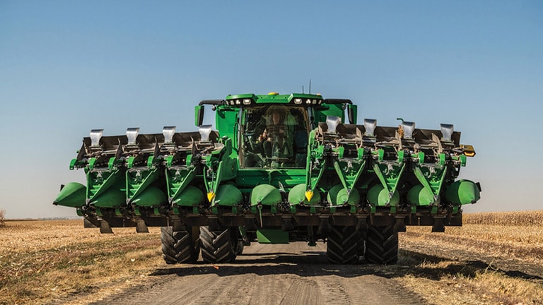 Combine harvester with multiple corn heads moves down a dirt path in a sunny agricultural field. (2nd thumbnail)