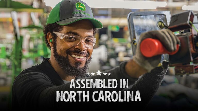 A smiling person safety glasses and a Deere hat assembling a piece of equipment at the factory in North Carolina. (2nd thumbnail)