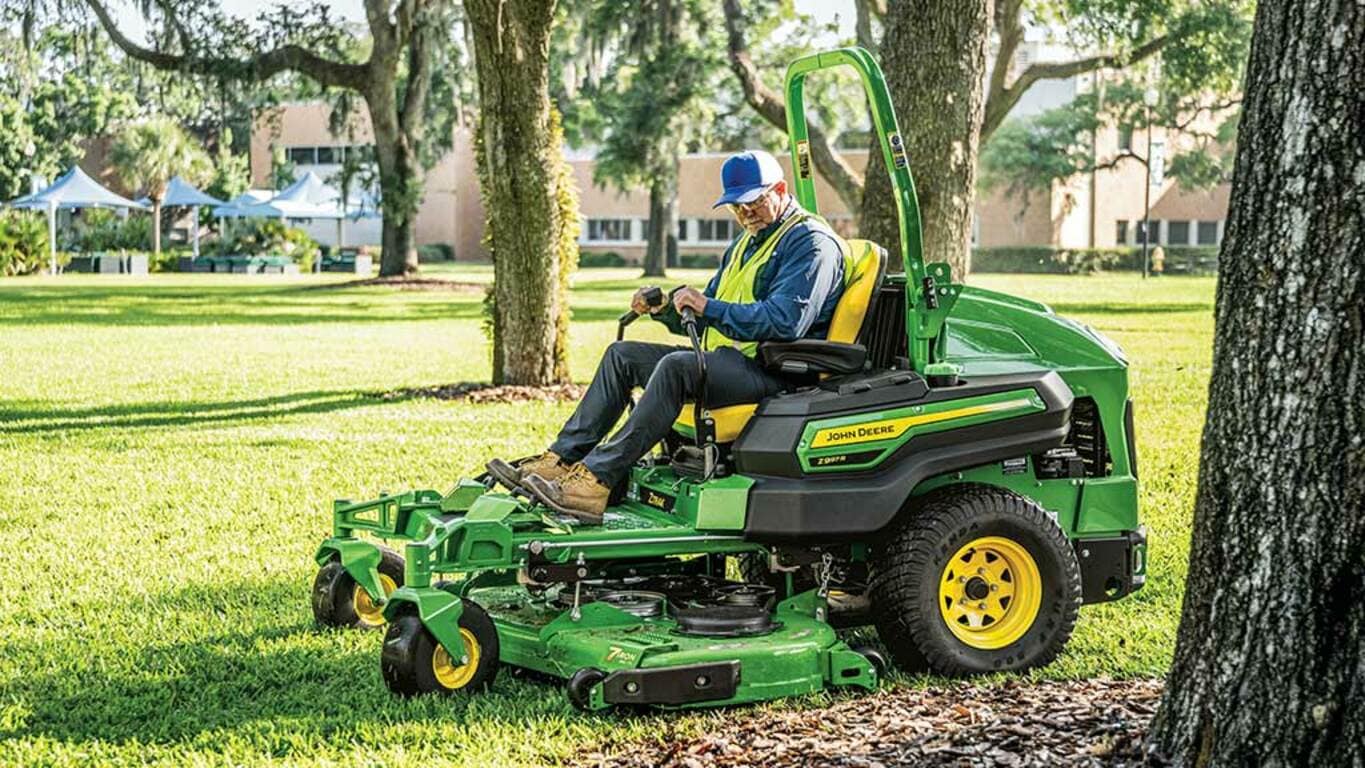 A person in a yellow work vest mowing a college campus lawn with a green zero-turn mower.