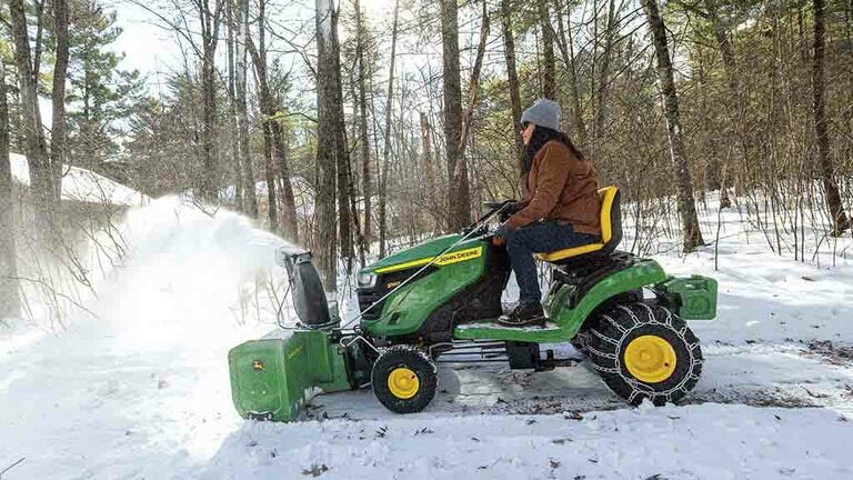 Snow being blown by a green riding mower equipped with a snow blower and snow chains. (10th thumbnail)
