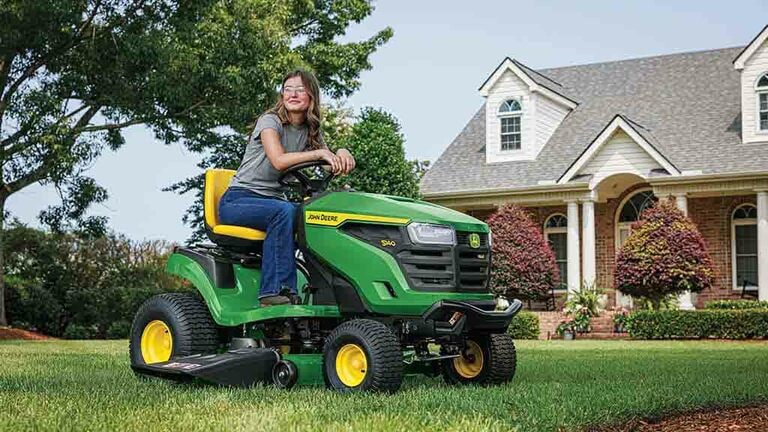 A smiling person resting on a green riding mower on a well-kept lawn. (11th thumbnail)