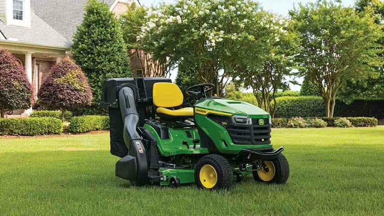 A green riding mower with a bagging attachment sitting on a lawn with short even grass. (7th thumbnail)
