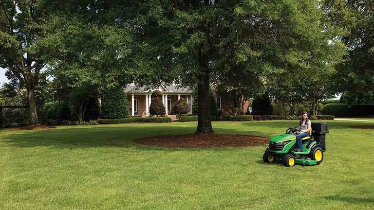A wide view of a well-kept lawn, off to the side a green riding mower with a bagging attachment. (6th thumbnail)