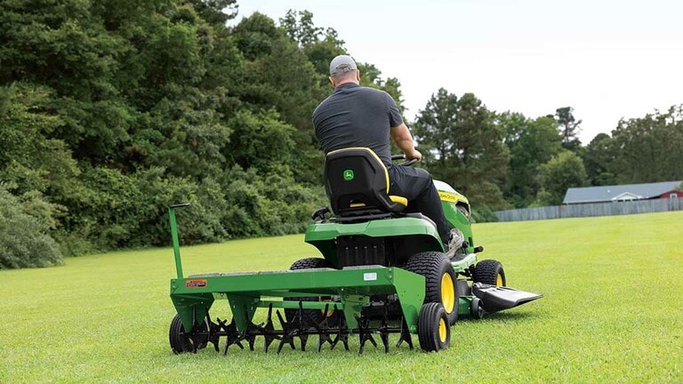 Rear view of a green lawn aerator being pulled by a green riding mower through an open grassy field. (6th thumbnail)