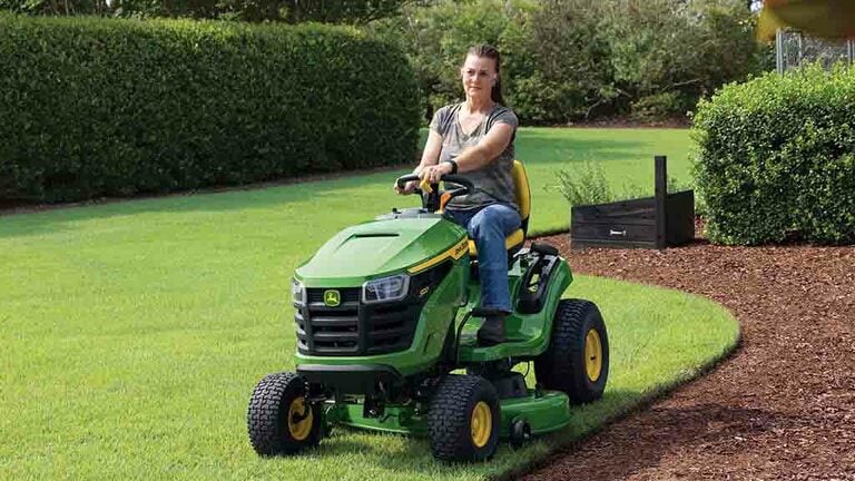 A person mowing the edge of a lawn by a patch of mulch on a green riding mower. (6th thumbnail)