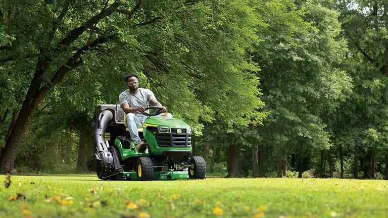 A person driving a green riding mower with a bagging attachment over a well-kept lawn. (11th thumbnail)