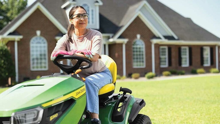 A person smiling while resting on a green riding mower with a on a well-kept lawn. (11th thumbnail)