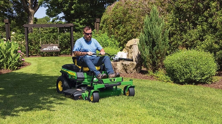 A person driving a green zero-turn mower around mulched areas with a swing, bushes, and rocks. (7th thumbnail)