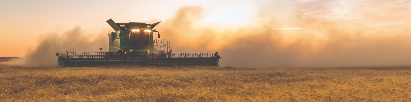 Front shot of a John Deere combine harvesting wheat at dusk, with LED lighting shining through dust.