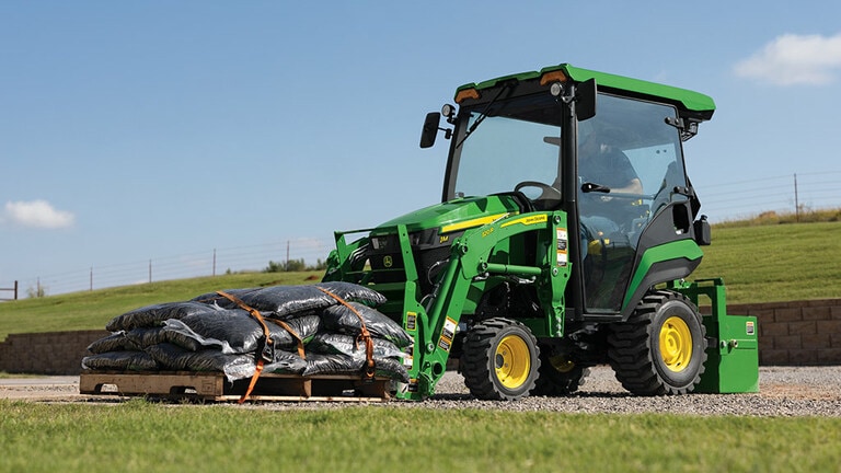 A pallet fork attached to a cabbed green compact tractor lifts a pallet of filled black bags. (7th thumbnail)