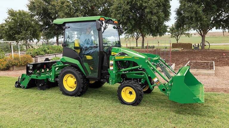 A green compact tractor with a loader and ground seeder drives next to a garden plot. (5th thumbnail)