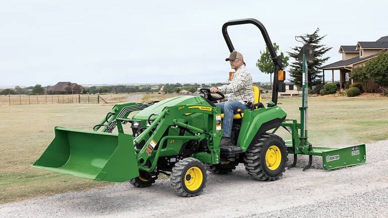 A person driving a green compact tractor equipped with a loader and land plane spreading gravel on a gravel road. (6th thumbnail)