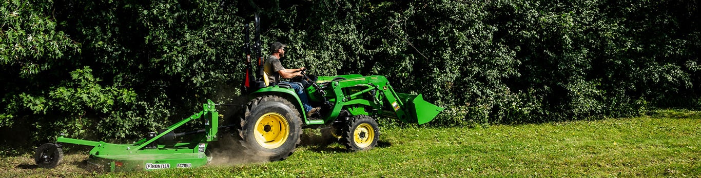 A rotary cutter is pulled by a green compact tractor cutting grass along a treeline.
