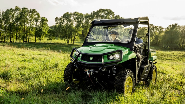 Two people in a green Gator drive in an open grassy field with thin trees in the background. (3rd thumbnail)