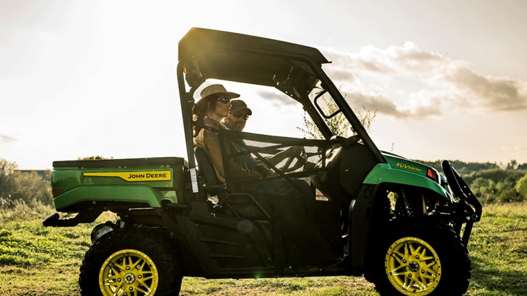 A Gator carrying two passengers off-roads in a field with the sun shining directly behind it. (4th thumbnail)