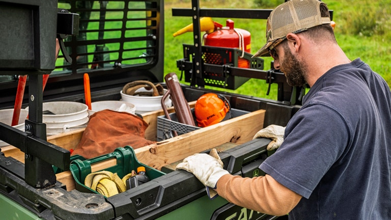 A person looks into a cargo box full of various tools and supplies on the back of an olive-green Gator. (13th thumbnail)