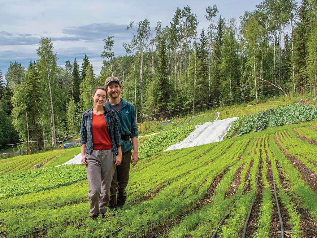 Two smiling farmers standing between rows of emerging crops in a field surrounded by trees