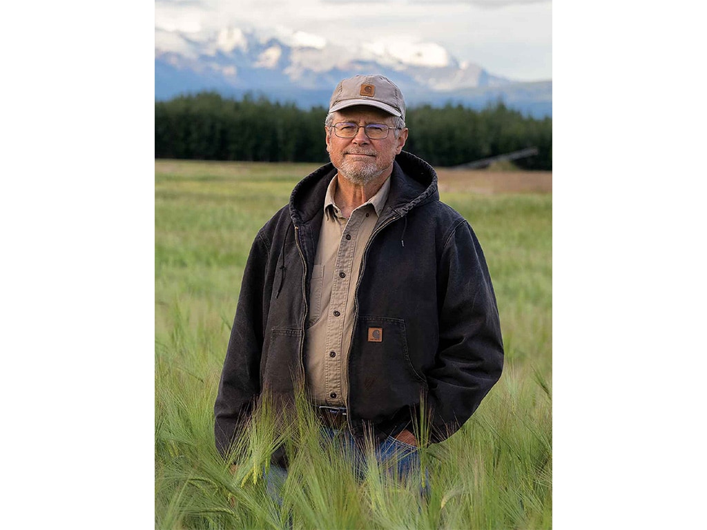 smiling farmer with baseball cap and glasses on standing in a wheat field with snowcapped mountains blurred in the distance