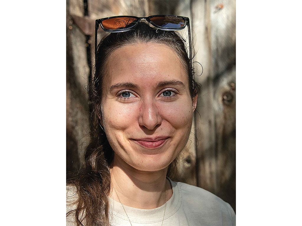 a smiling young person with long dark brown hair, sunglasses atop their head, wearing a white tshirt