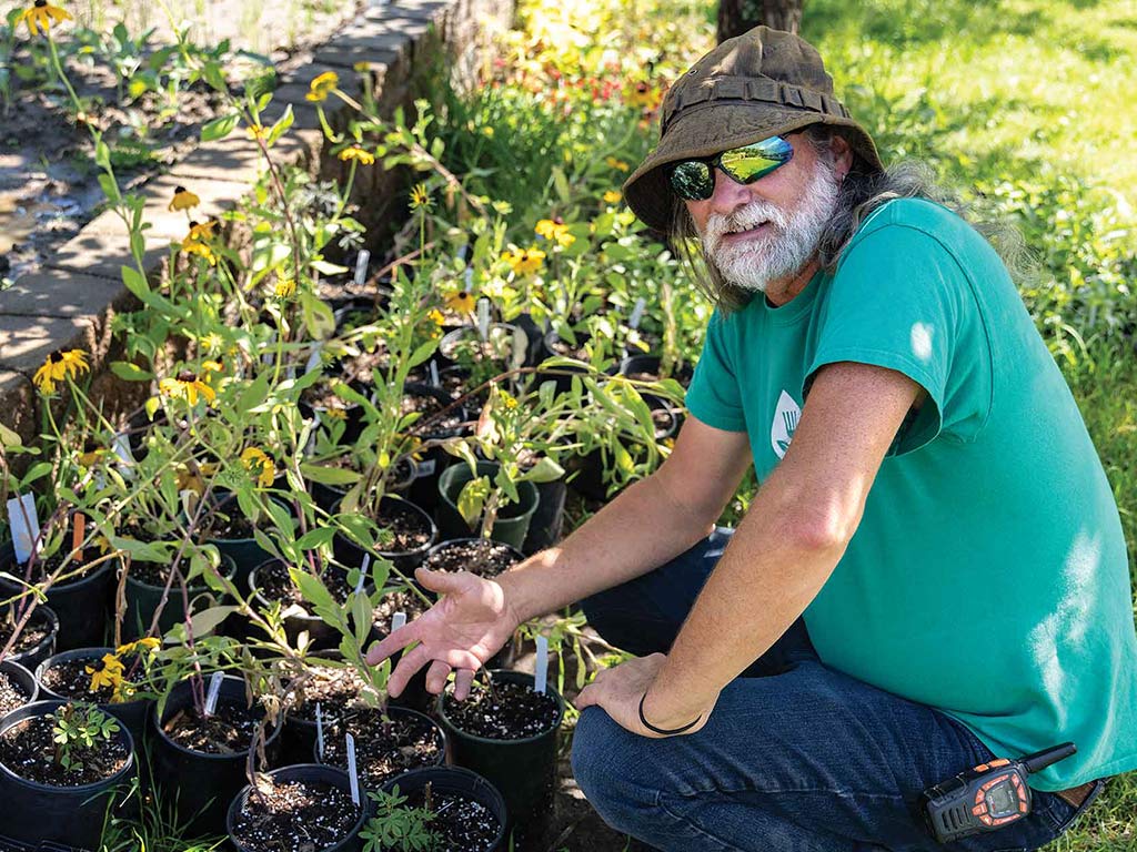 smiling volunteer with white beard, wrap-around sunglasses and fishing hat sitting in front of plants in black plastic planters