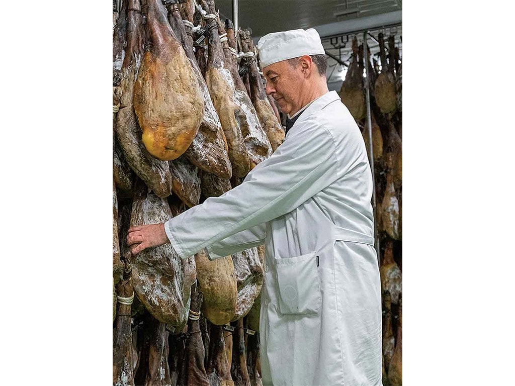 a butcher in a white coat and chef's hat inspecting aging legs of ham