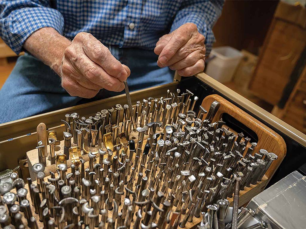 organized set of leather craftsman tools on a wooden work bench with the worker's hands in view