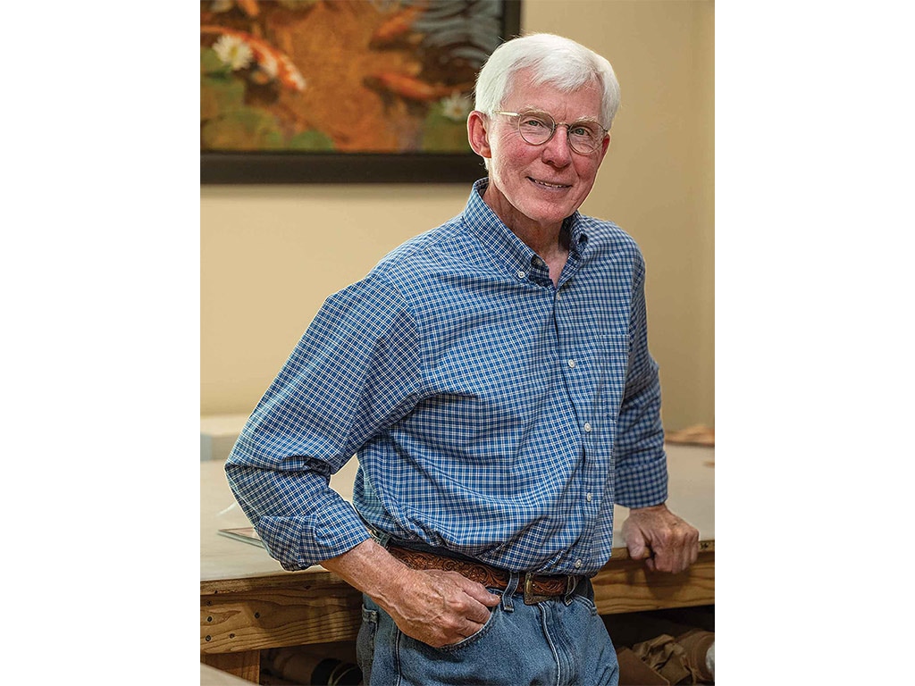 a smiling person with white hair and glasses wearing a blue button-down shirt and jeans