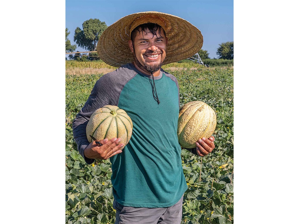 a smiling farmer cradling a melon in each arm with a wide brimmed straw hat standing in a field