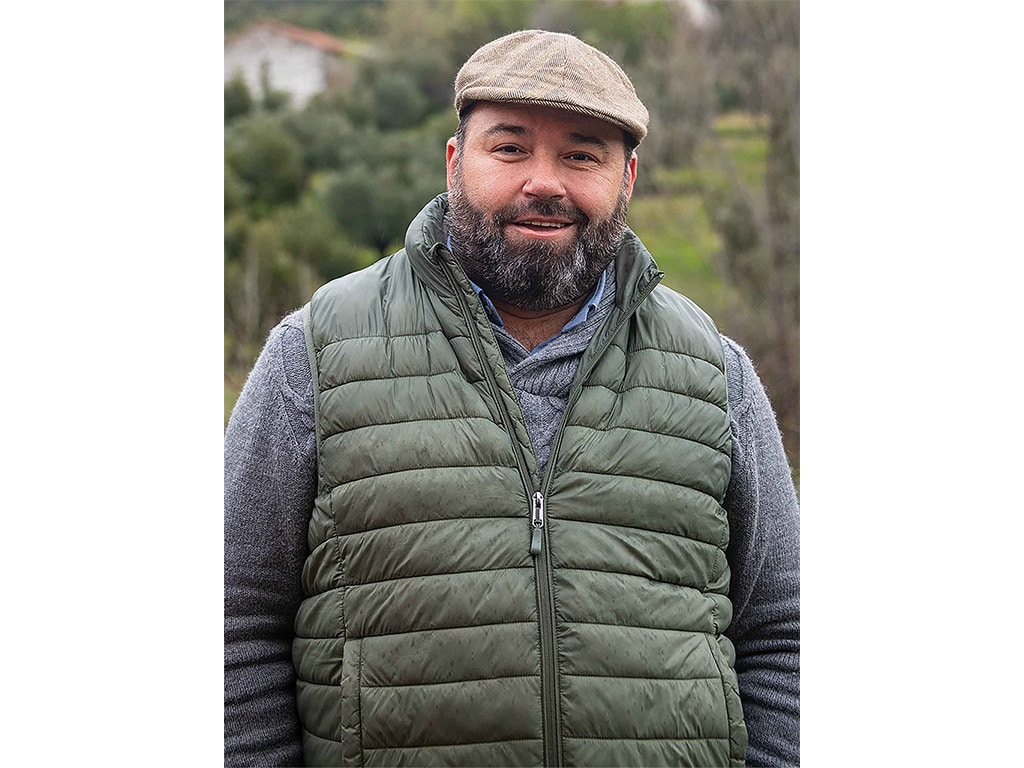 a smiling olive farmer with a beard and derby cap wearing an olive green puffer vest