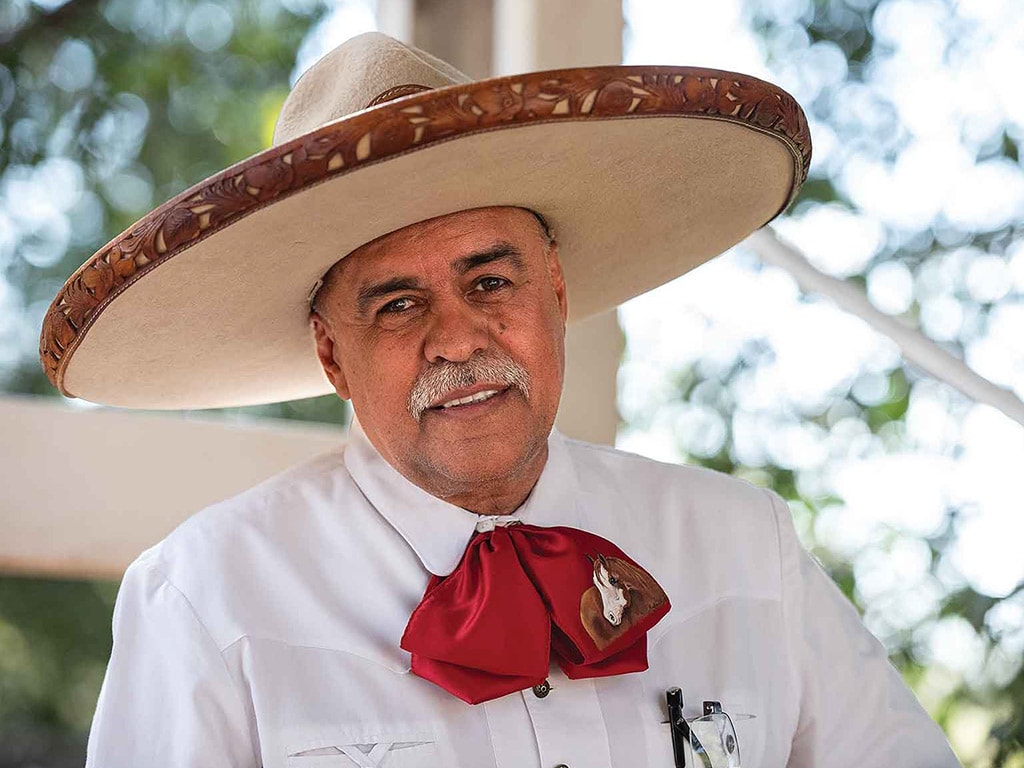 the escaramuza announcer adorned with a white collared shirt, satin red bow tie with a horse embroidered on it, and a cream-colored wide brimmed hat with an ornate leather edge