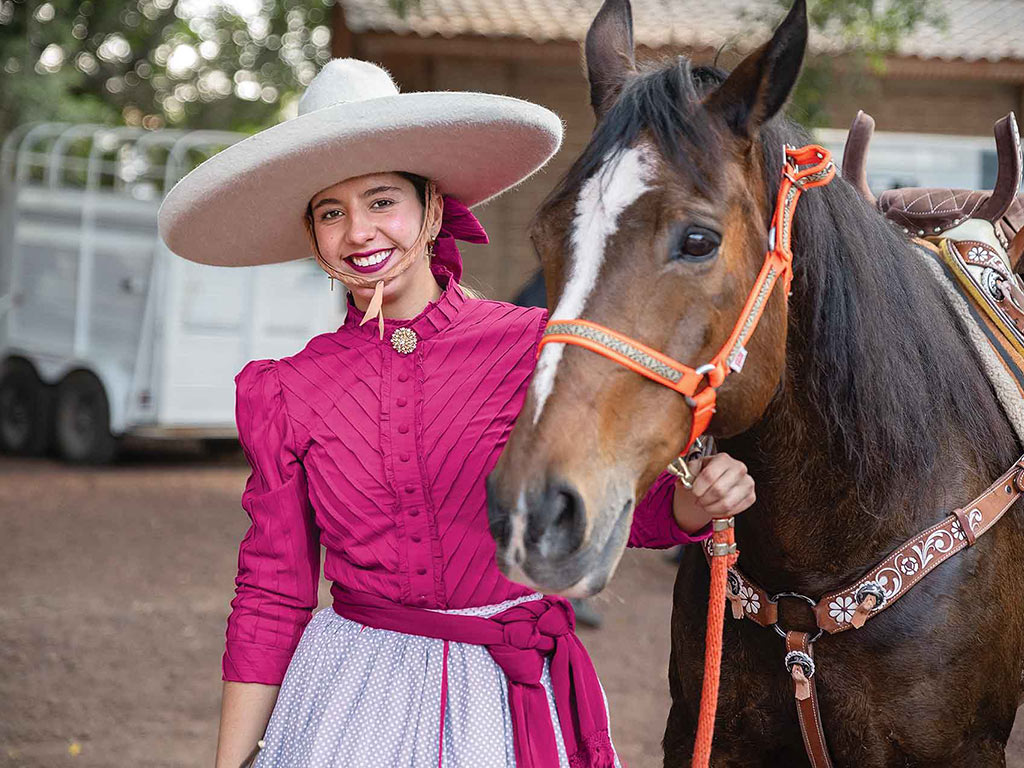 a smiling escaramuza rider wearing a bright fuchsia coat and bone white wide brimmed hat with a rounded crown holding the bridle of their horse