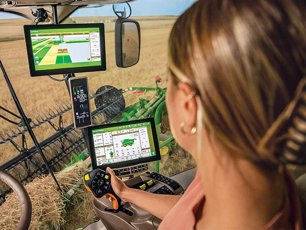 Interior of tractor cab with three small monitors and a farmer handling a joystick while harvesting a grain field